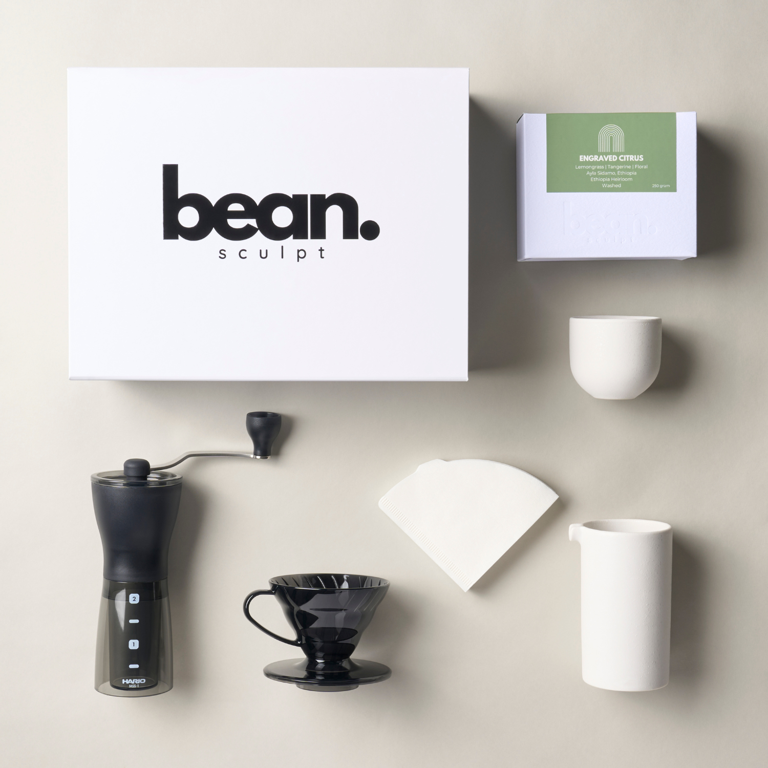 Flat-lay of a Coffee Starter Bundle. Includes a Hario hand grinder, V60 matte black dripper, Bean Sculpt specialty coffee, 300ml jug, and flat white coffee cup on a white surface.