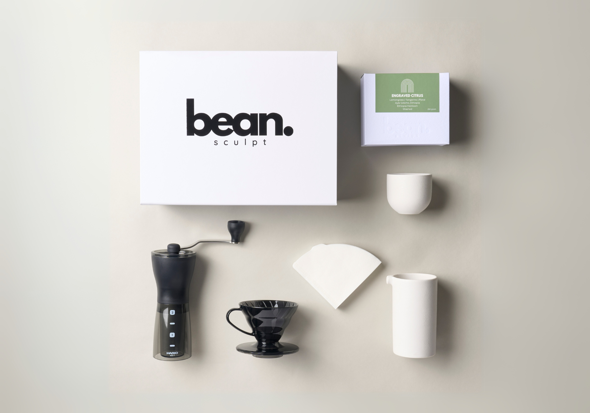 Flat-lay of a coffee brewing set featuring a Hario coffee grinder, Hario V60 dripper, coffee server, white coffee cup, and a 250g bag of Bean Sculpt specialty coffee, arranged on a clean surface.